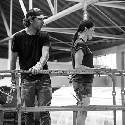 Black and white photo of founders, Ilana and Sterling, on scissor lift in TMB production area