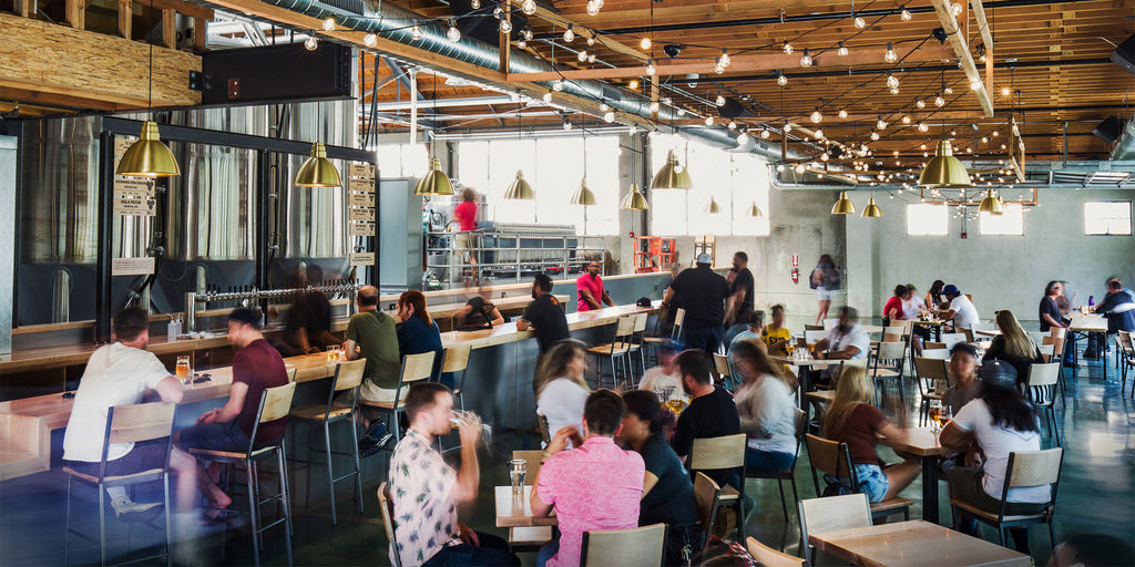 Photo of crowded taproom interior with sparkling festoon lights and many people enjoying Trademark Brewing beers