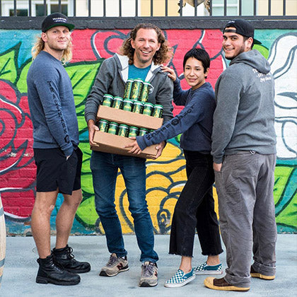 Photo of four Trademark Brewing employees holding cases of canned beer with colorful mural in the background