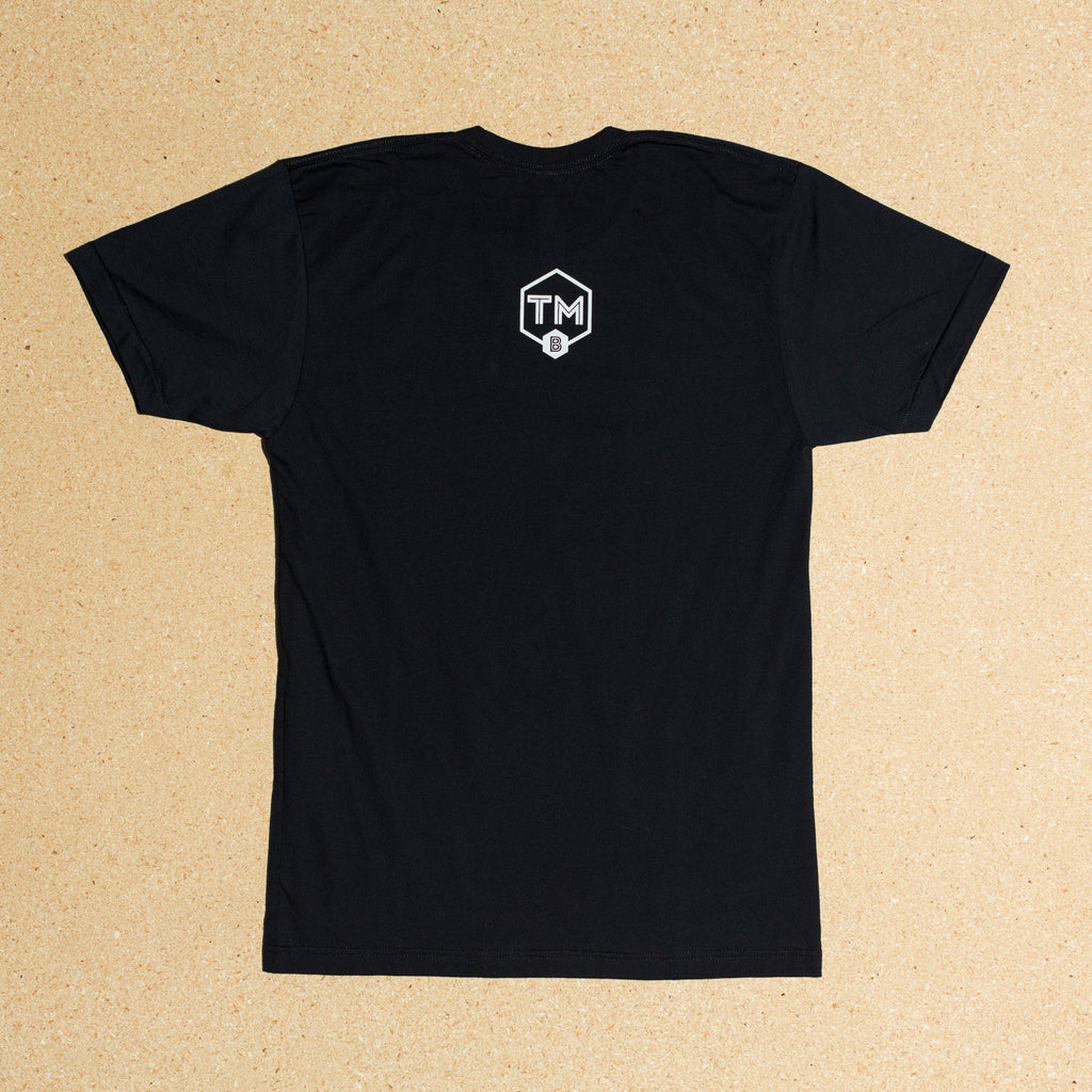 Photo of black t-shirt with large Trademark Brewing small shield logo on back side